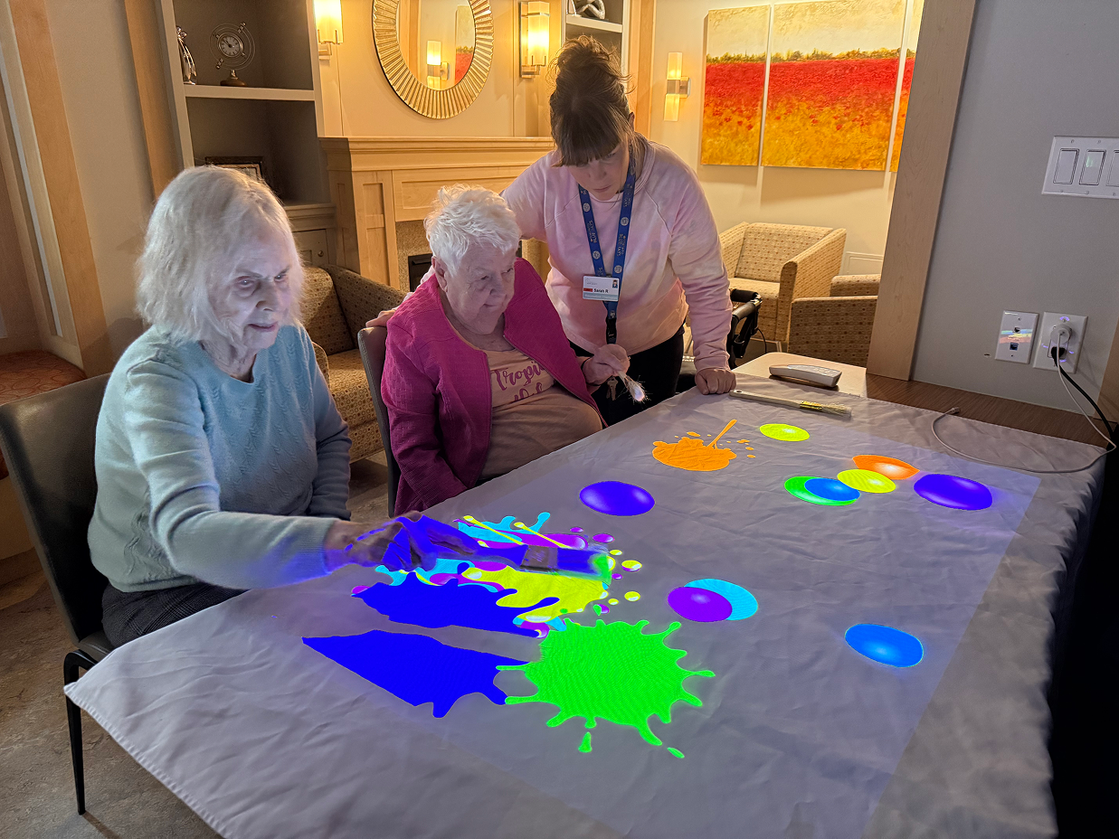 Two elderly women and a caregiver interacting with a colorful digital art projection on a table.
