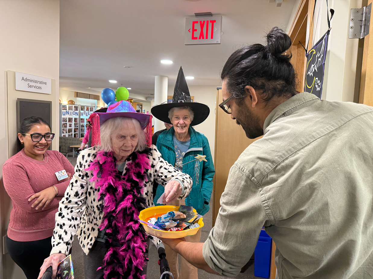 Elderly woman wearing a festive hat and feather boa takes candy from a bowl held by a man with a bun, while two women, one in a witch hat, smile nearby in a decorated hallway.