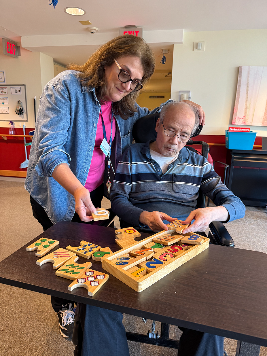 Caregiver assisting an elderly man in a wheelchair with a wooden number puzzle in a room with warm lighting.
