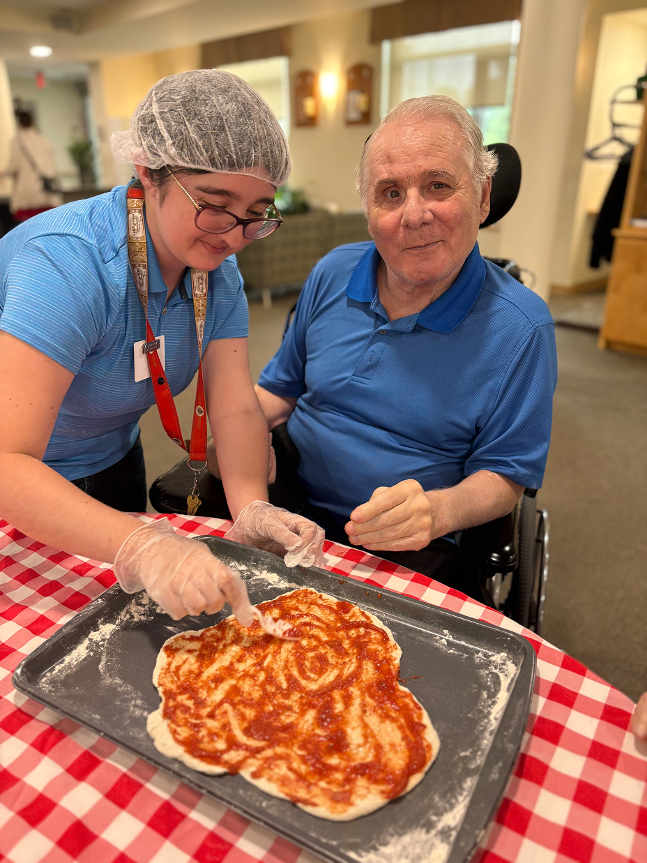 A woman in a hairnet and gloves spreading tomato sauce on pizza dough while a man in a wheelchair watches.