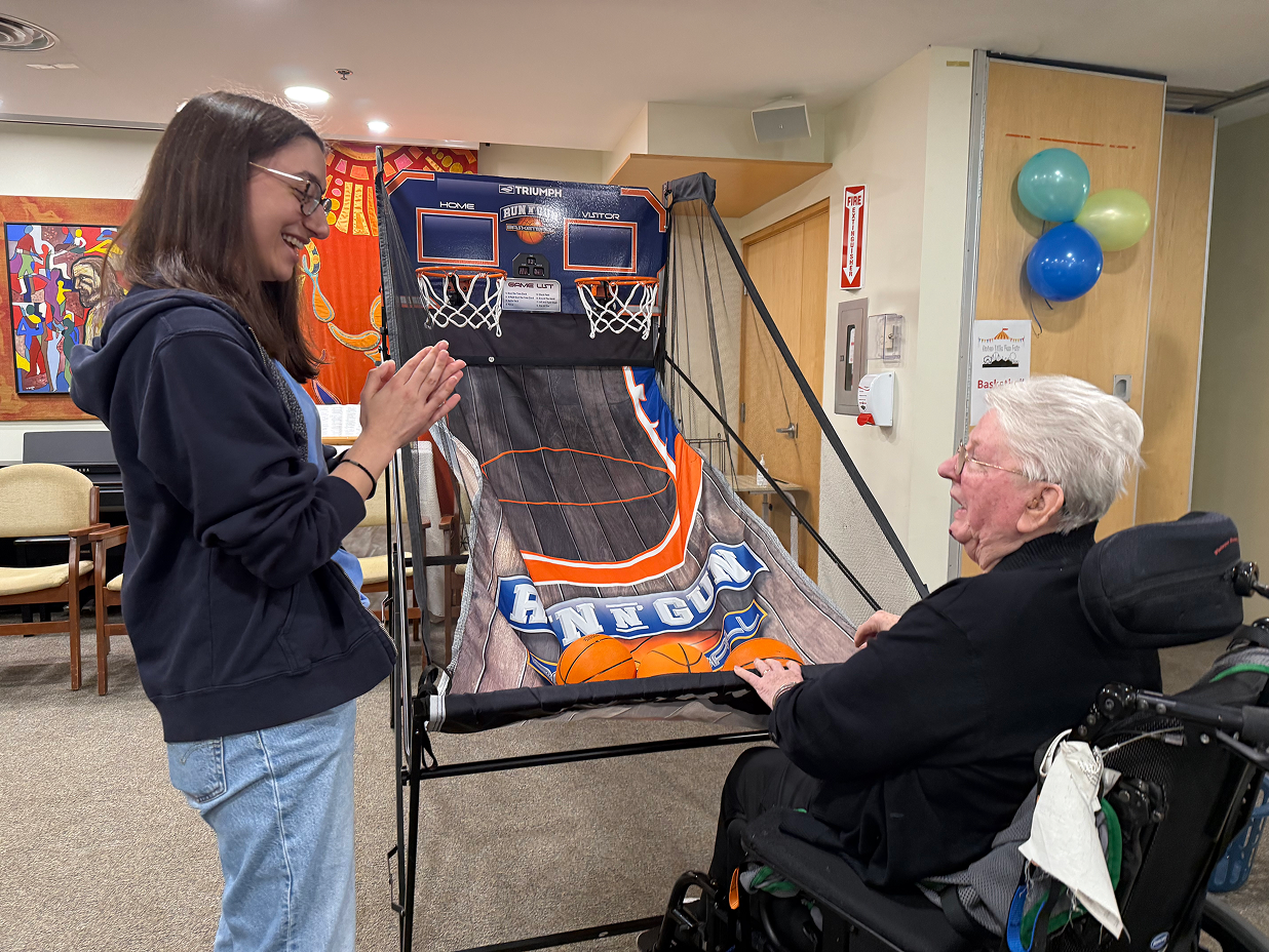 Young woman smiling and clapping hands while elderly person in a wheelchair plays indoor basketball arcade game.