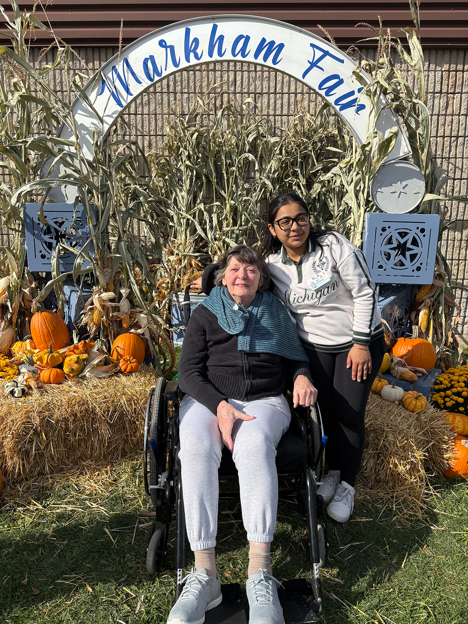 An elderly woman in a wheelchair and a younger woman standing next to her at a Markham Fair autumn display with pumpkins, corn stalks, and hay bales.
