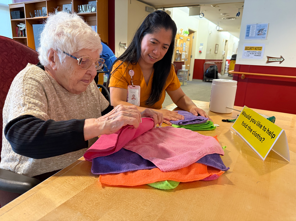 An elderly woman folds colorful cloths at a table with a caregiver assisting her in a communal room.