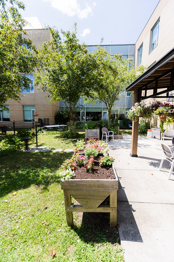 Sunny garden courtyard with raised wooden planters filled with colorful flowers, trees, and outdoor chairs near a building.