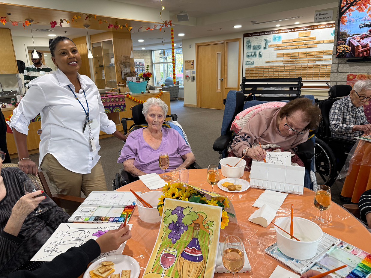 Group of elderly women in wheelchairs painting and socializing at a decorated table in a community center with a caregiver standing nearby.