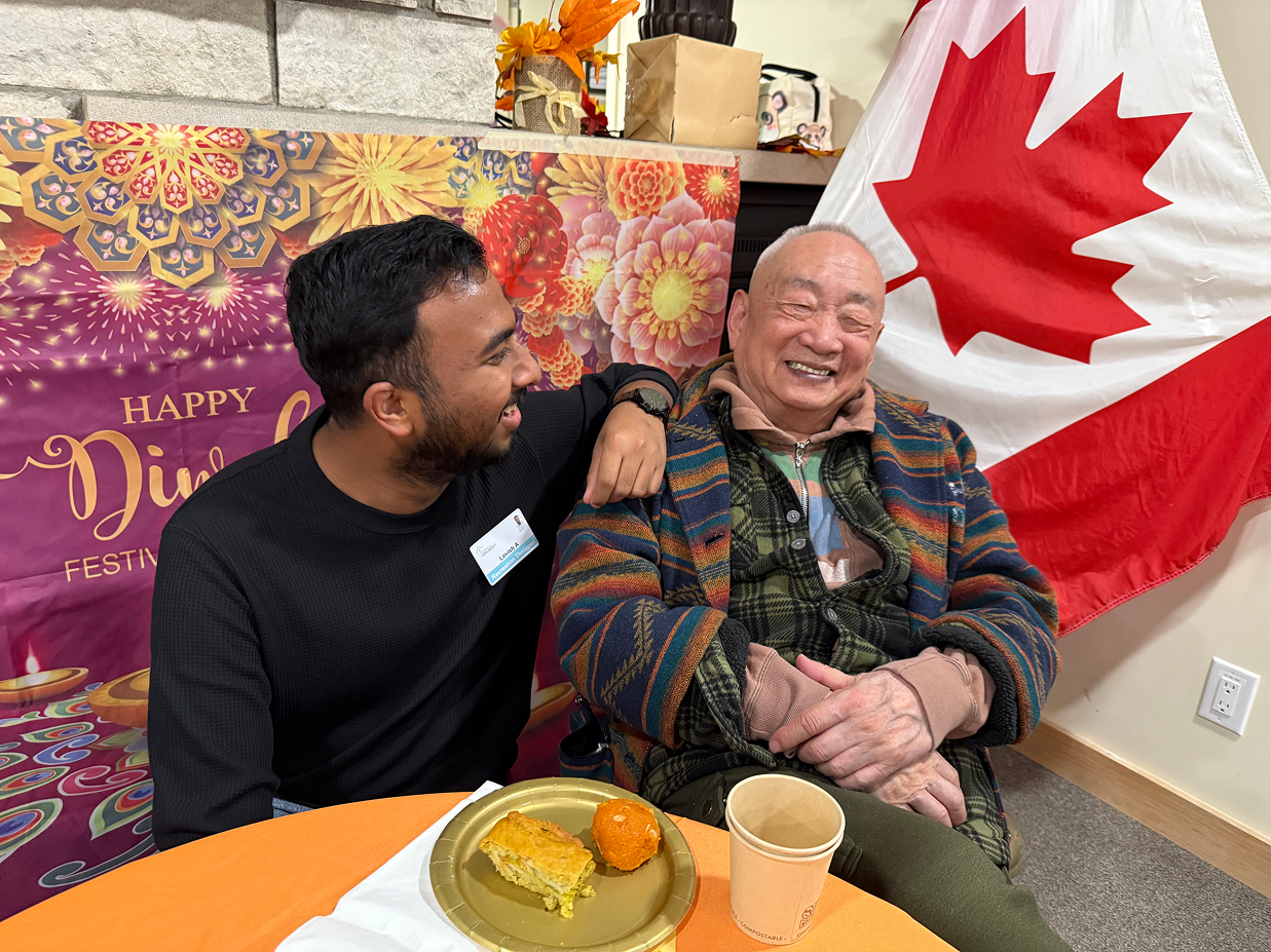 Two men smiling together at a Diwali festival table with Indian sweets and a Canadian flag in the background.