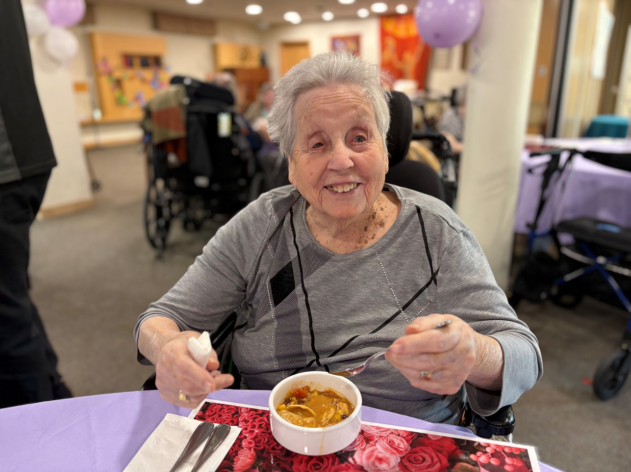 Smiling elderly woman sitting at a table with a bowl of soup, holding a spoon and napkin.