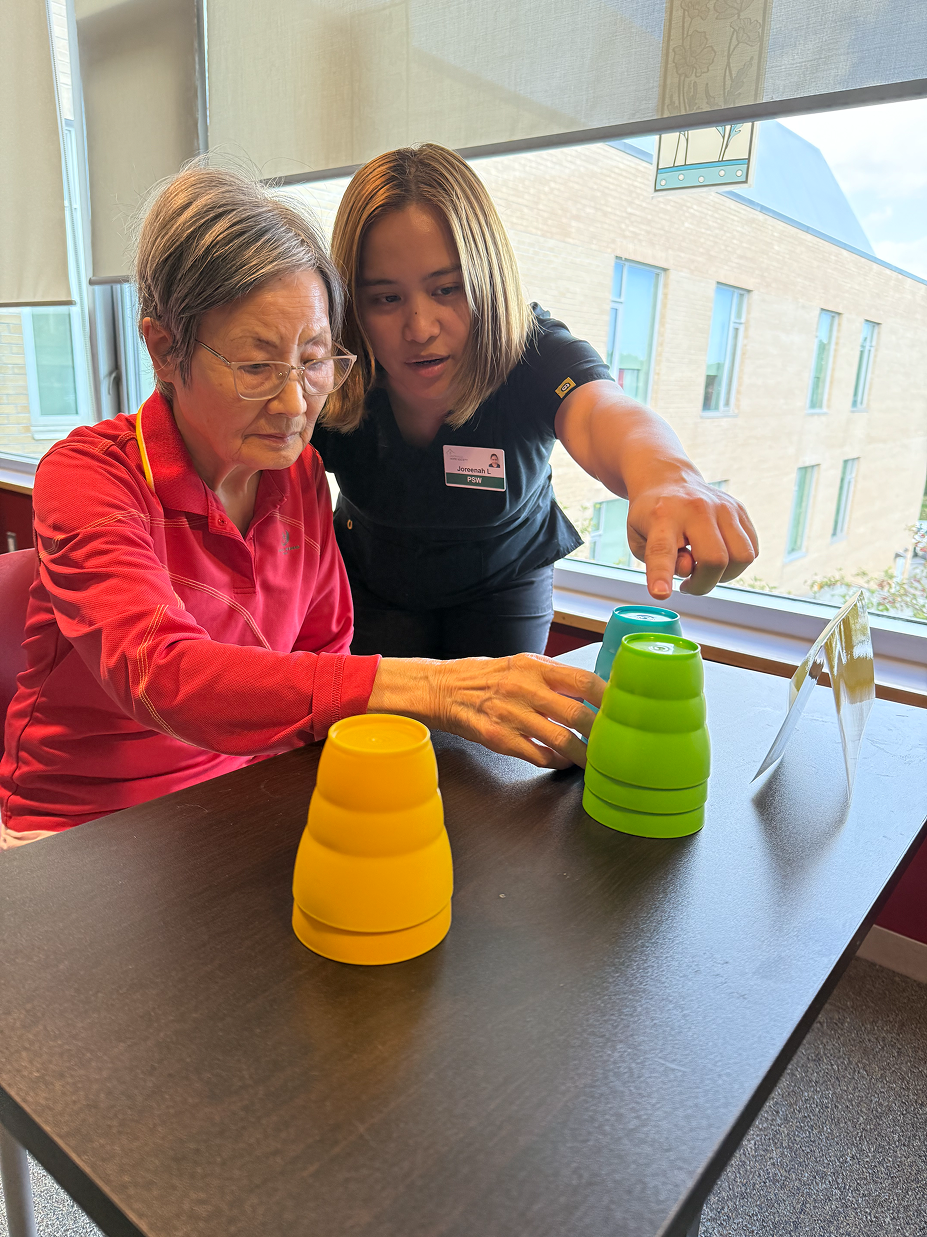 Caregiver guiding an elderly woman as they stack colorful plastic cups on a table near a window.