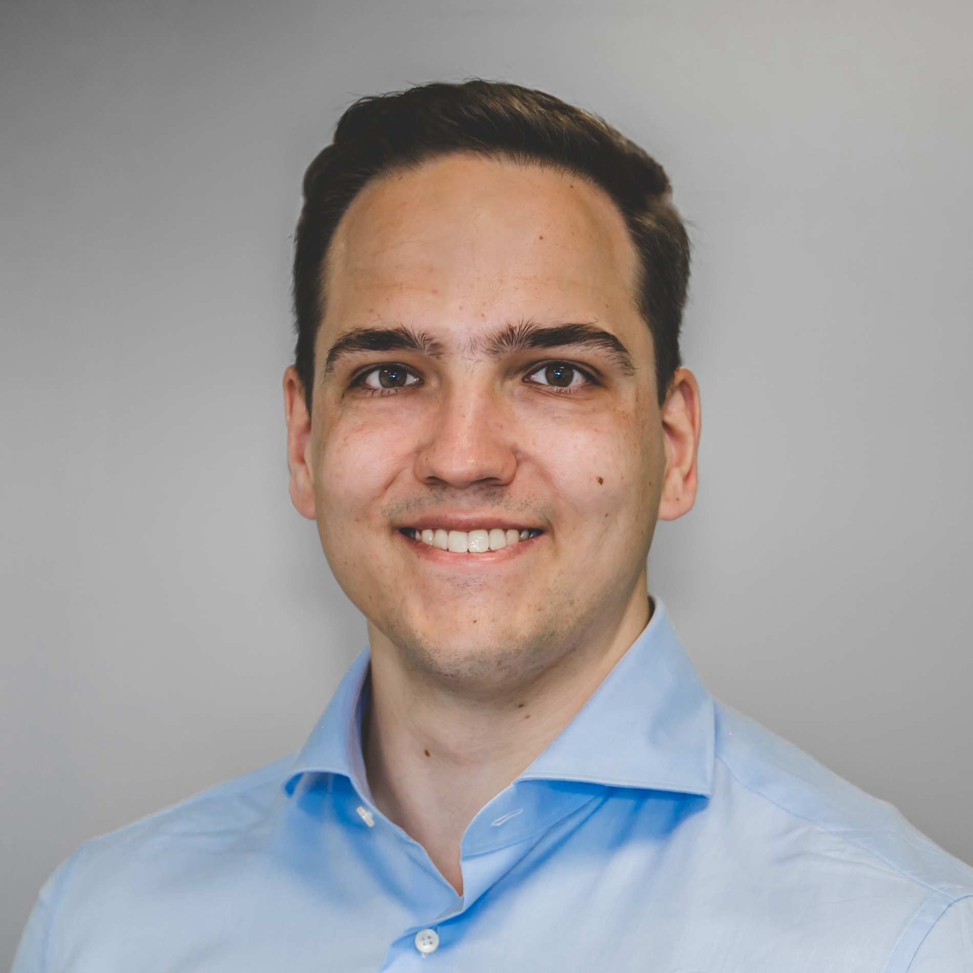 Portret van een lachende man met donker haar, een lichtblauw overhemd en een neutrale achtergrond.