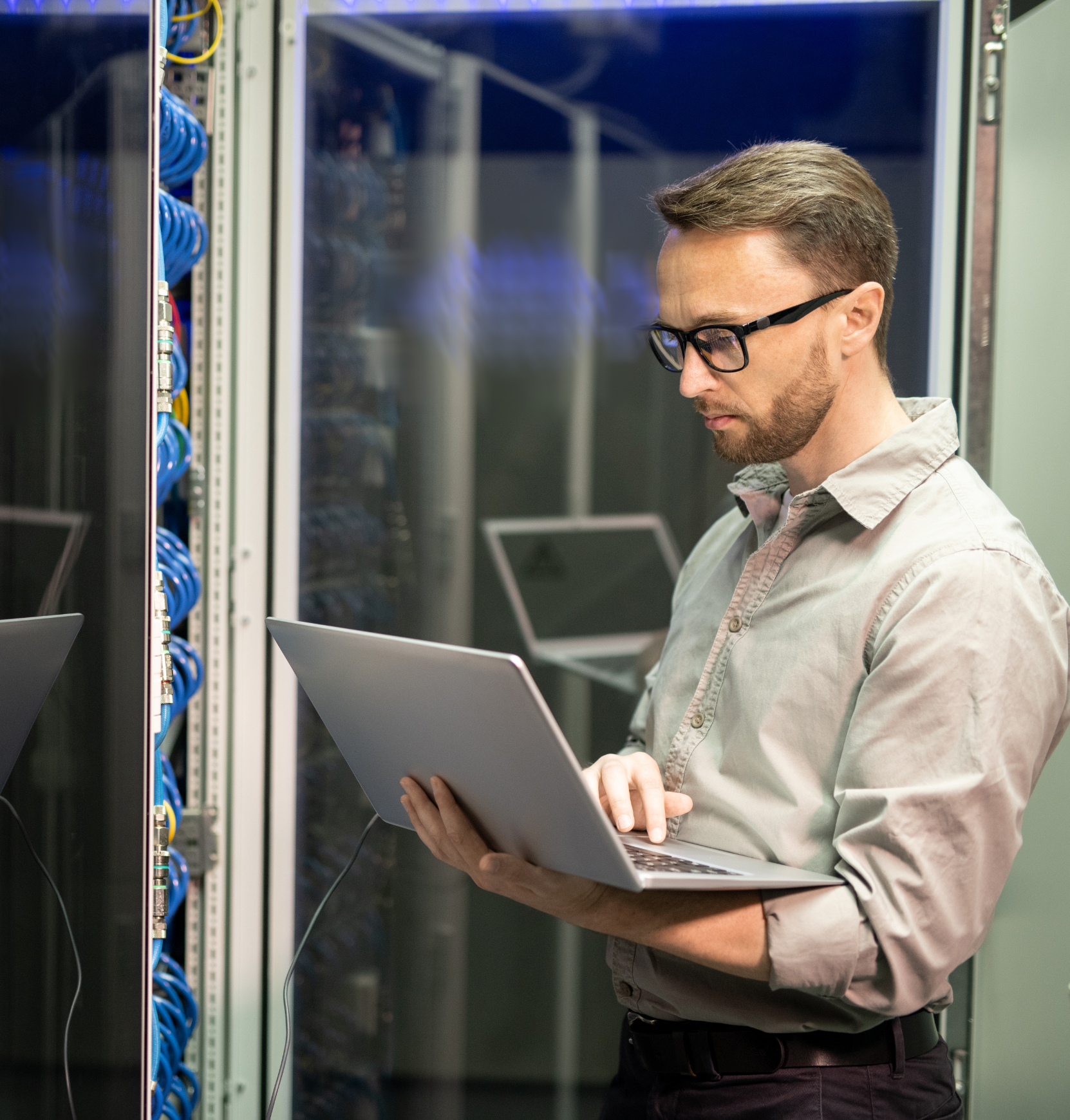 Man met bril controleert een laptop in een serverruimte naast netwerkbekabeling.