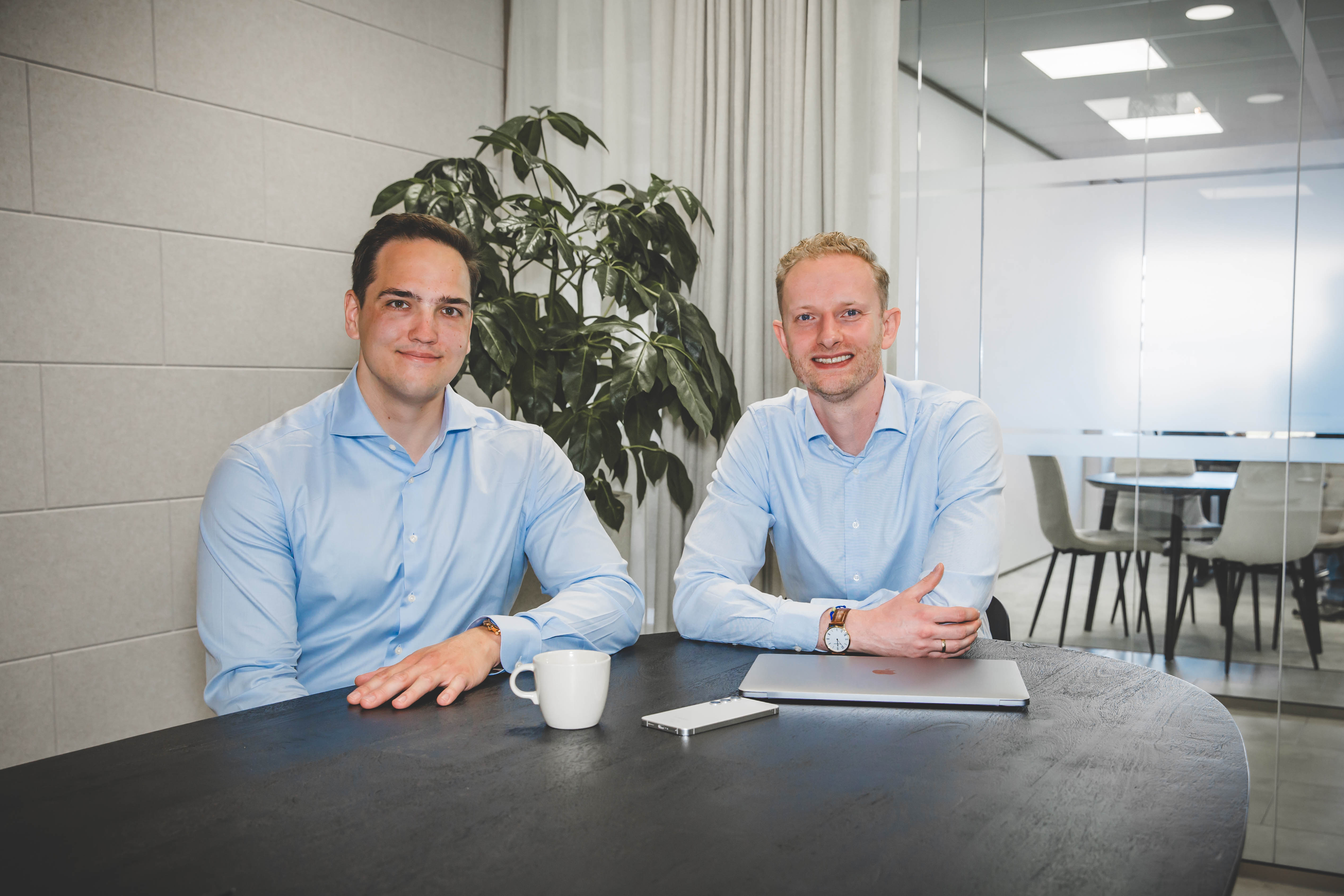 Twee mannen in lichtblauwe hemden zitten aan een tafel met een witte koffiekop, een smartphone en een gesloten laptop in een moderne kantoorruimte.