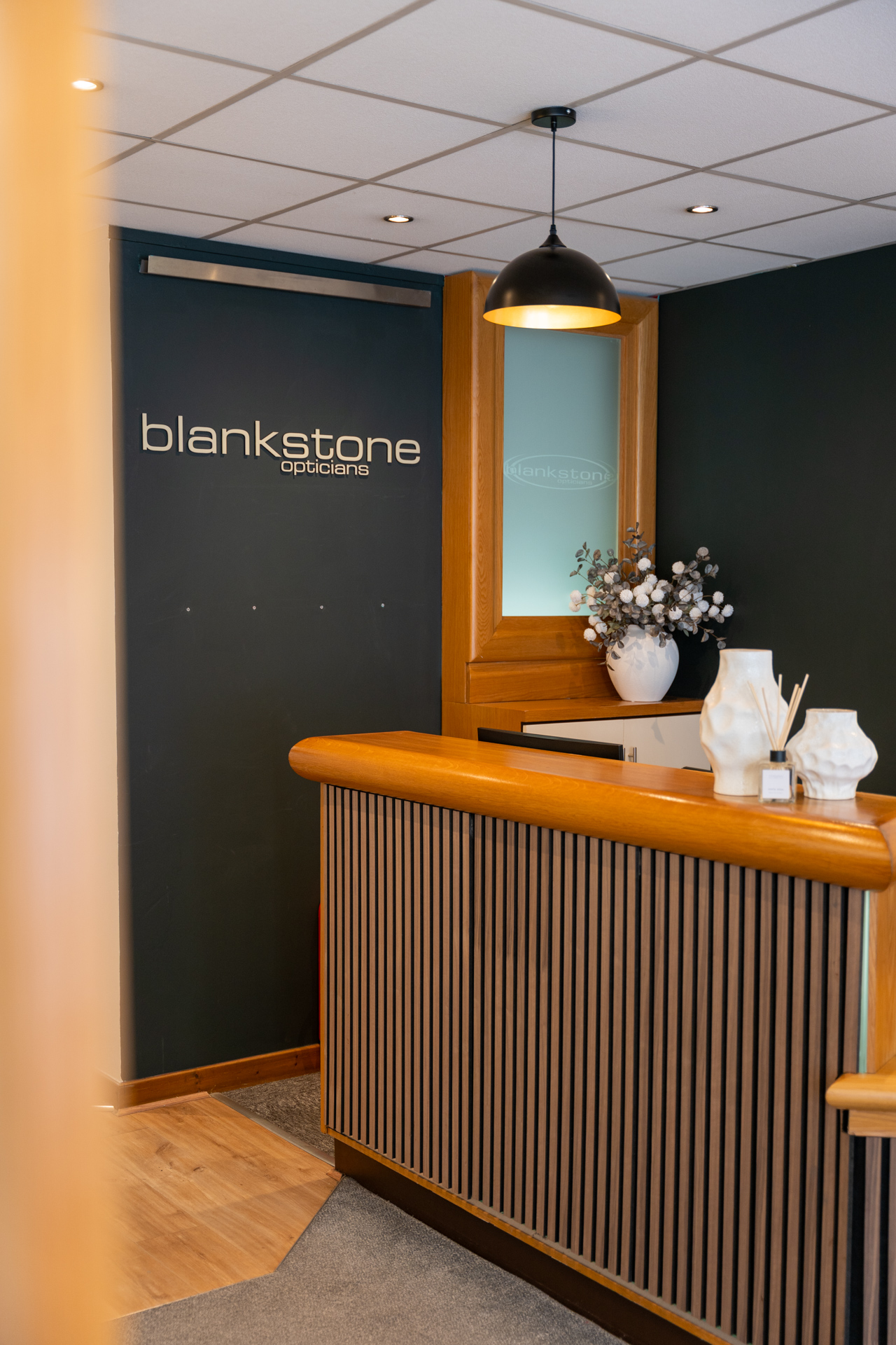 Modern Blankstone Opticians reception desk with wooden counter, pendant lamp, and dark signage.