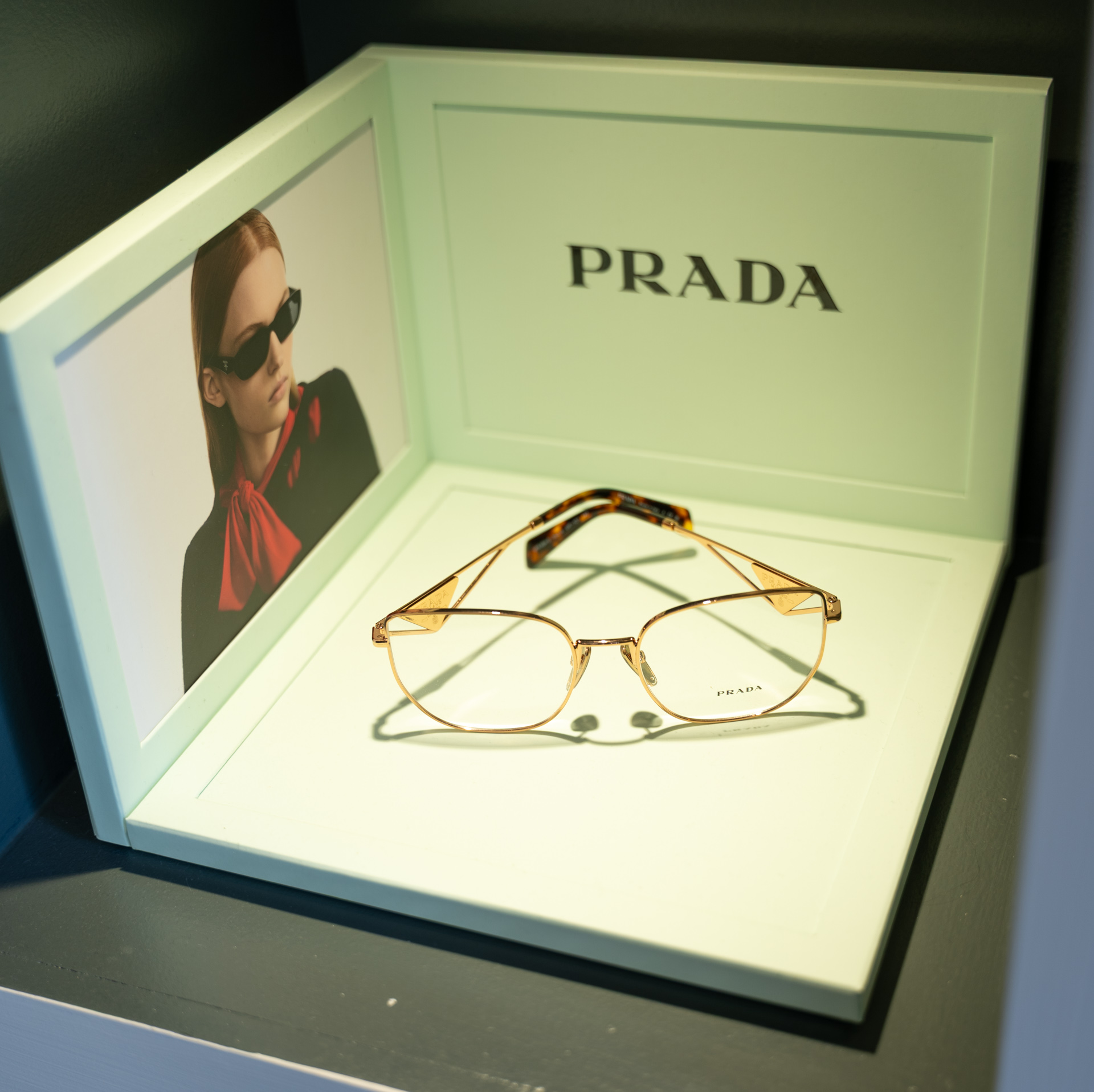 Prada luxury eyewear display box with gold-framed glasses and sunglasses advertisement