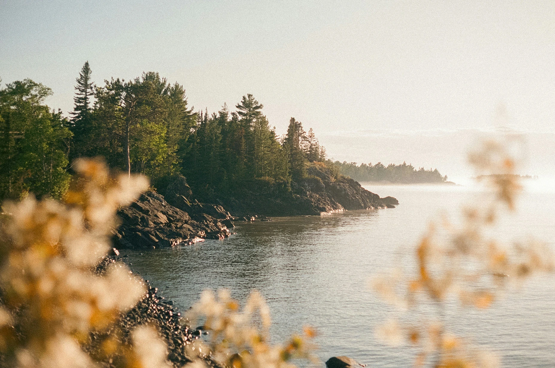 Paysage naturel au lever du soleil sur les rives rocheuses d’un lac du Canada, entouré de forêts de pins et de sapins. L’eau calme reflète la lumière dorée, créant une atmosphère paisible et sauvage. Photo d’un itinéraire de road trip en van au Canada avec Beaux Périples