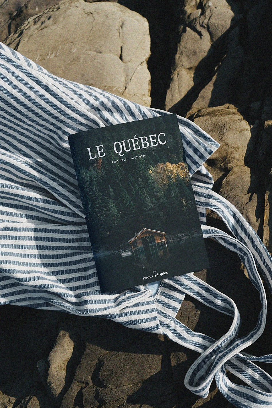 Carnet de voyage personnalisé “Le Québec” par Beaux Périples, posé sur un tissu rayé en extérieur. Road trip sur mesure au Canada incluant forêts, lacs et découvertes en famille