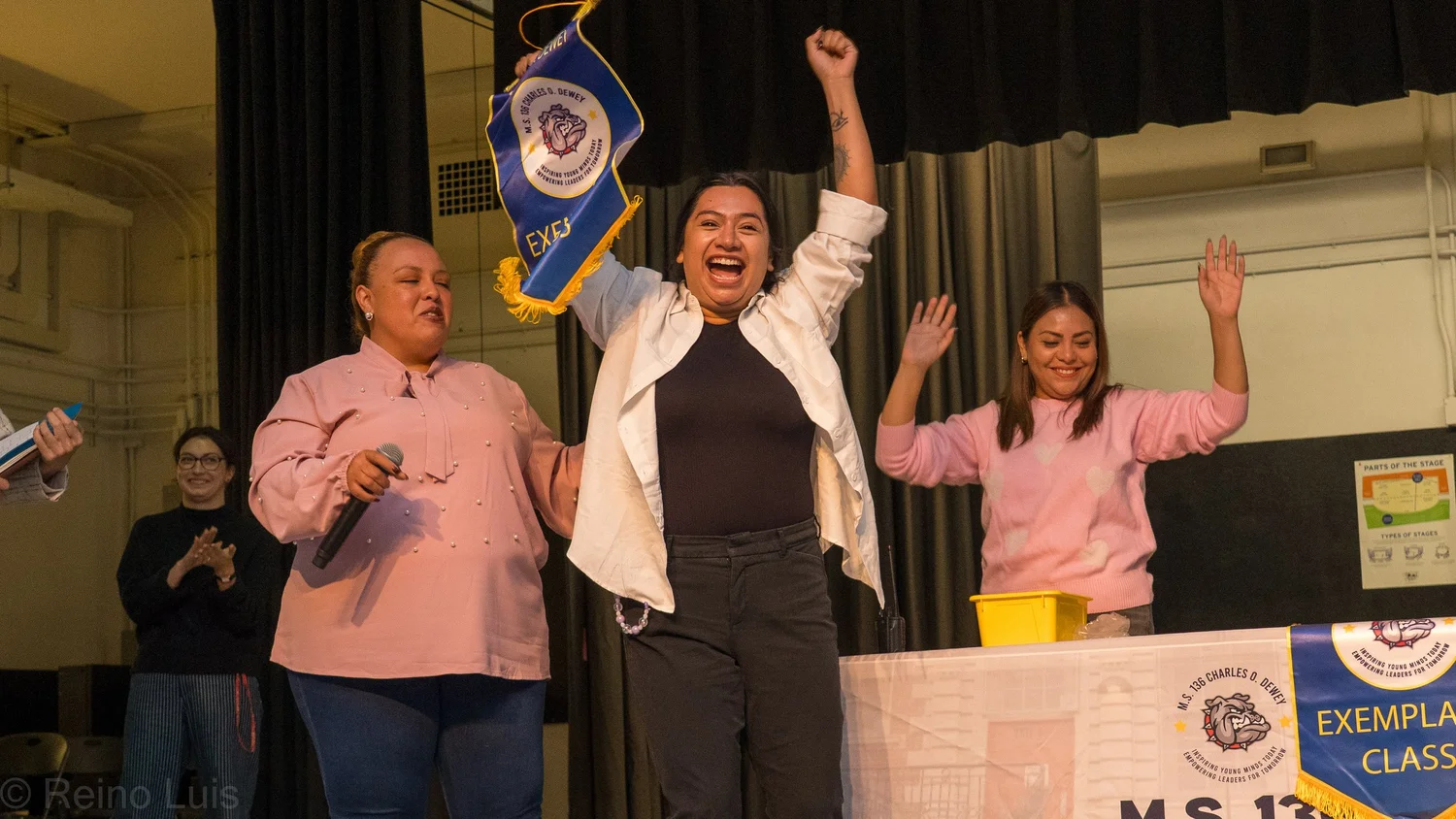 Three adults celebrate on stage at MS 136 awards ceremony