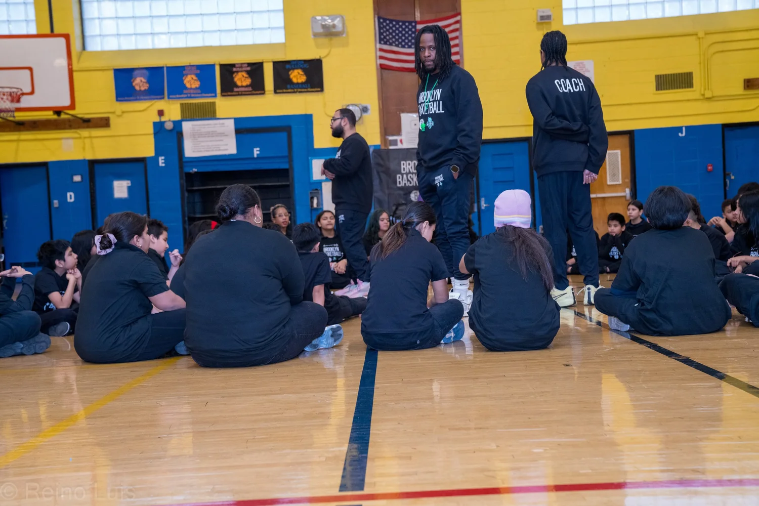 Brooklyn Basketball coaches addressing students seated on gymnasium floor