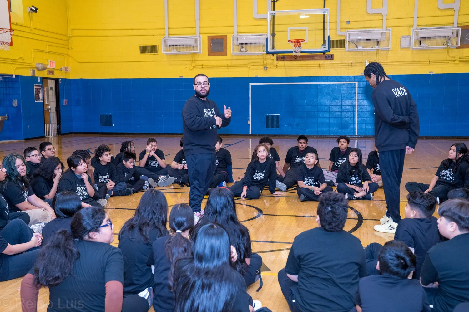 Brooklyn Nets coaches addressing students seated on gymnasium floor