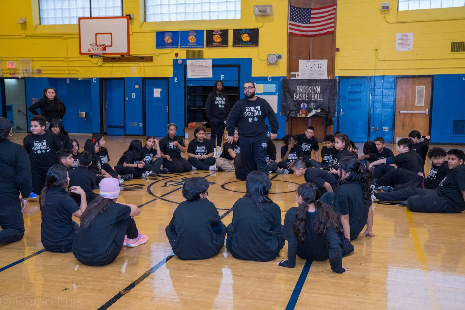 Brooklyn Basketball coach addresses students seated on gymnasium floor