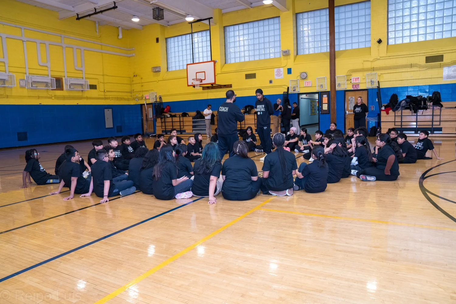 Coach addresses students seated on gymnasium floor during Brooklyn Basketball visit