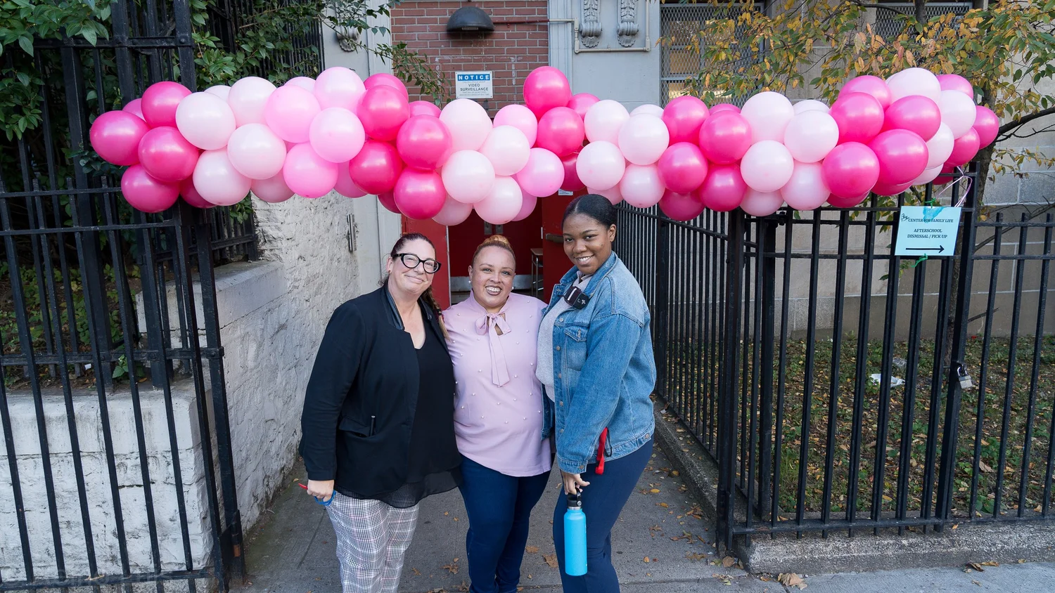 Three staff members posing under pink balloon arch at school entrance