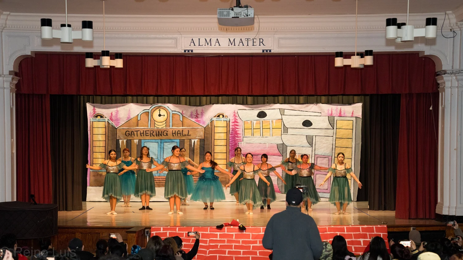 Students in teal costumes perform a dance on school auditorium stage