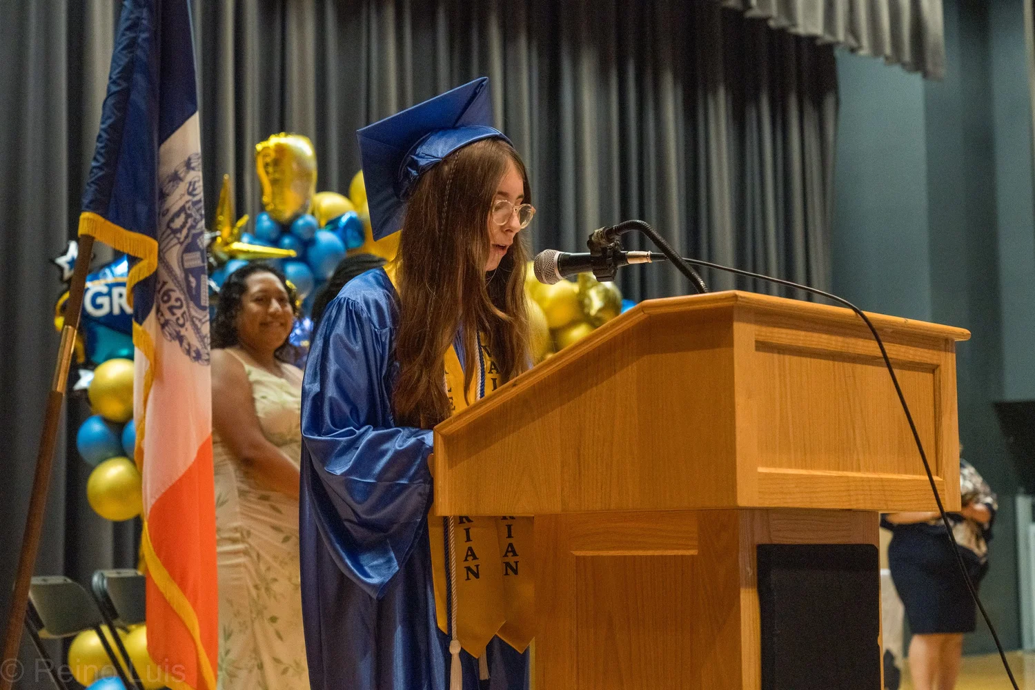 Graduate in blue cap and gown speaking at graduation podium