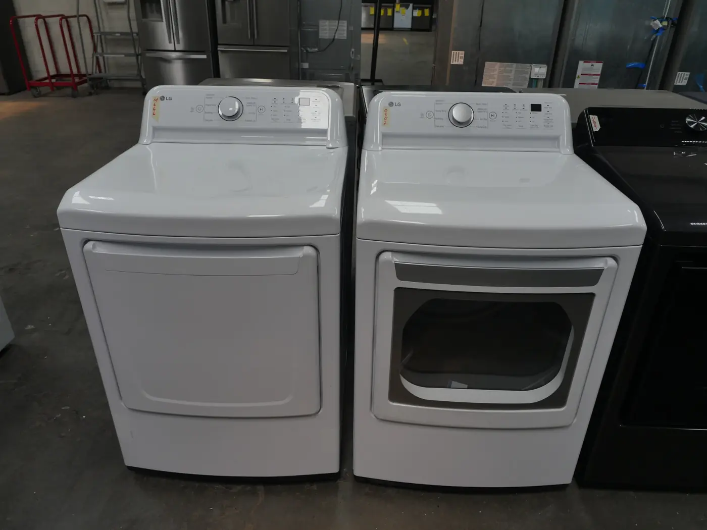 Two white LG laundry machines side by side in a showroom, the left is a dryer with a solid front door and the right is a dryer with a glass front door, both with control knobs and buttons on the top panel.