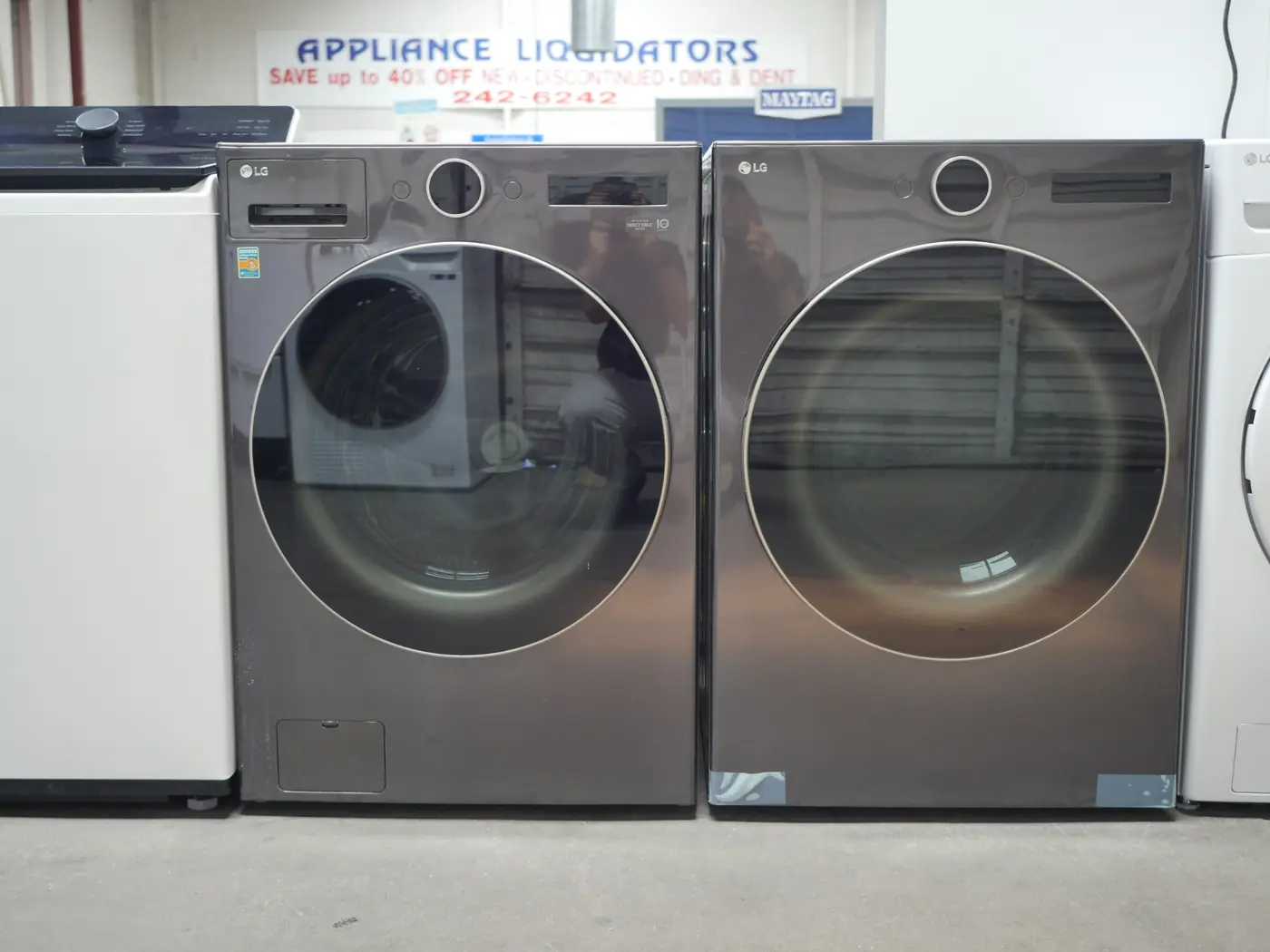 Pair of front-load LG washer and dryer units in a graphite finish displayed side by side on a showroom floor, with other white appliances visible beside them and a store sign in the background.