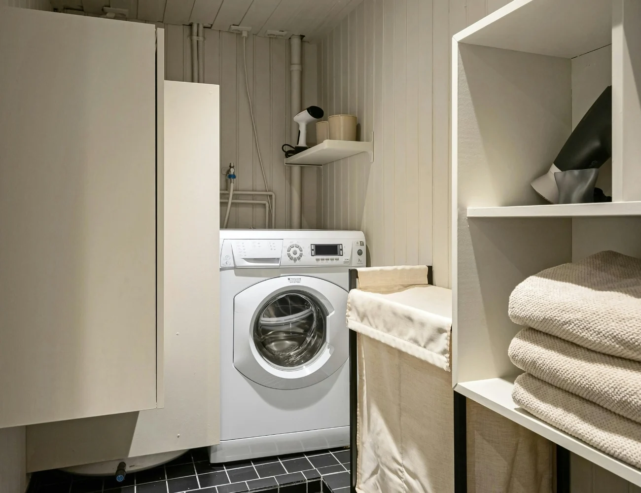 Small laundry room with a white front-loading washing machine, a beige fabric laundry hamper, wall shelf holding a handheld steamer and container, and a white shelving unit with folded towels and a black hair dryer.