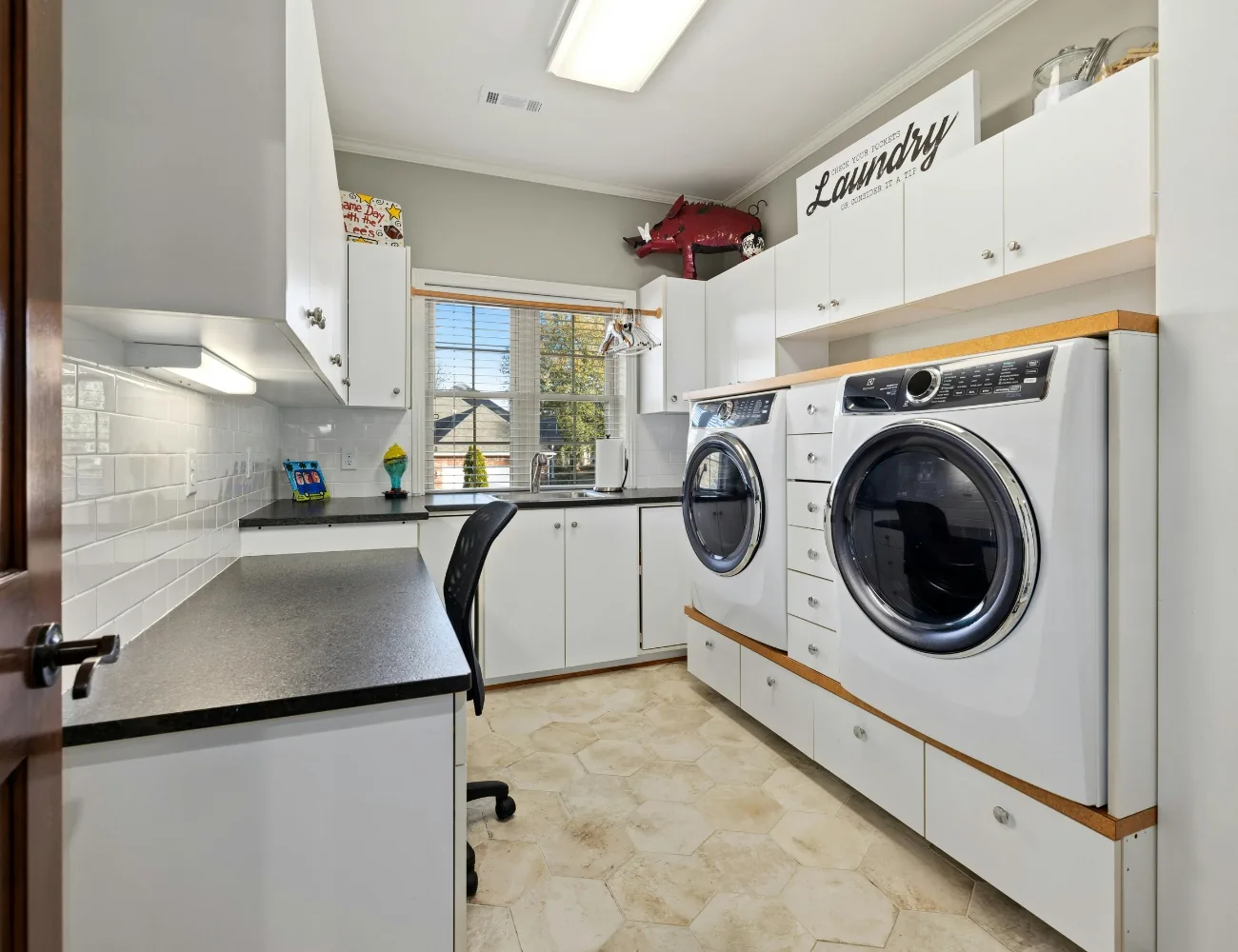 Bright laundry room with white cabinetry, black countertops, a front-loading washing machine and dryer, a window with blinds, a black office chair, and decorative items including a red pig sculpture and a sign reading 'Laundry'.