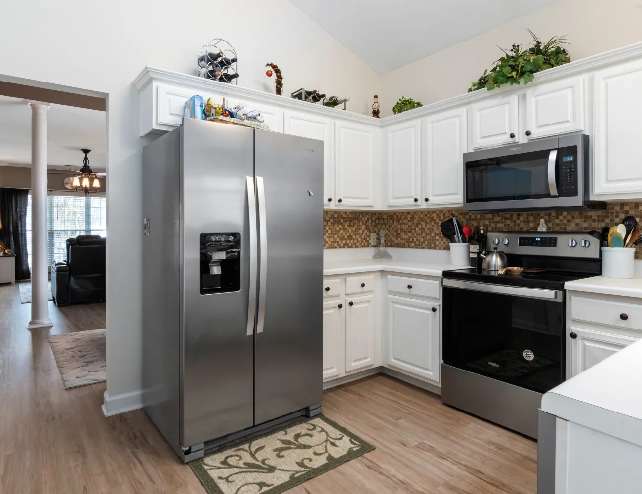 Modern kitchen corner featuring a stainless steel side-by-side refrigerator with an ice and water dispenser, white cabinetry with dark knobs, a matching stainless steel oven with a stovetop and an overhead microwave, mosaic tile backsplash, wooden floor, and decorative greenery on top of cabinets.