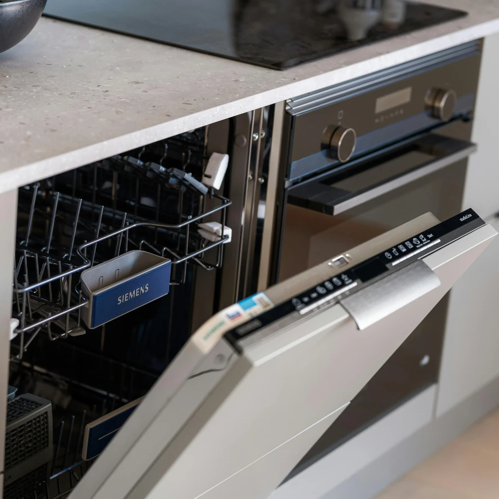 Open Siemens dishwasher installed under kitchen countertop next to a built-in oven, showing empty racks inside.