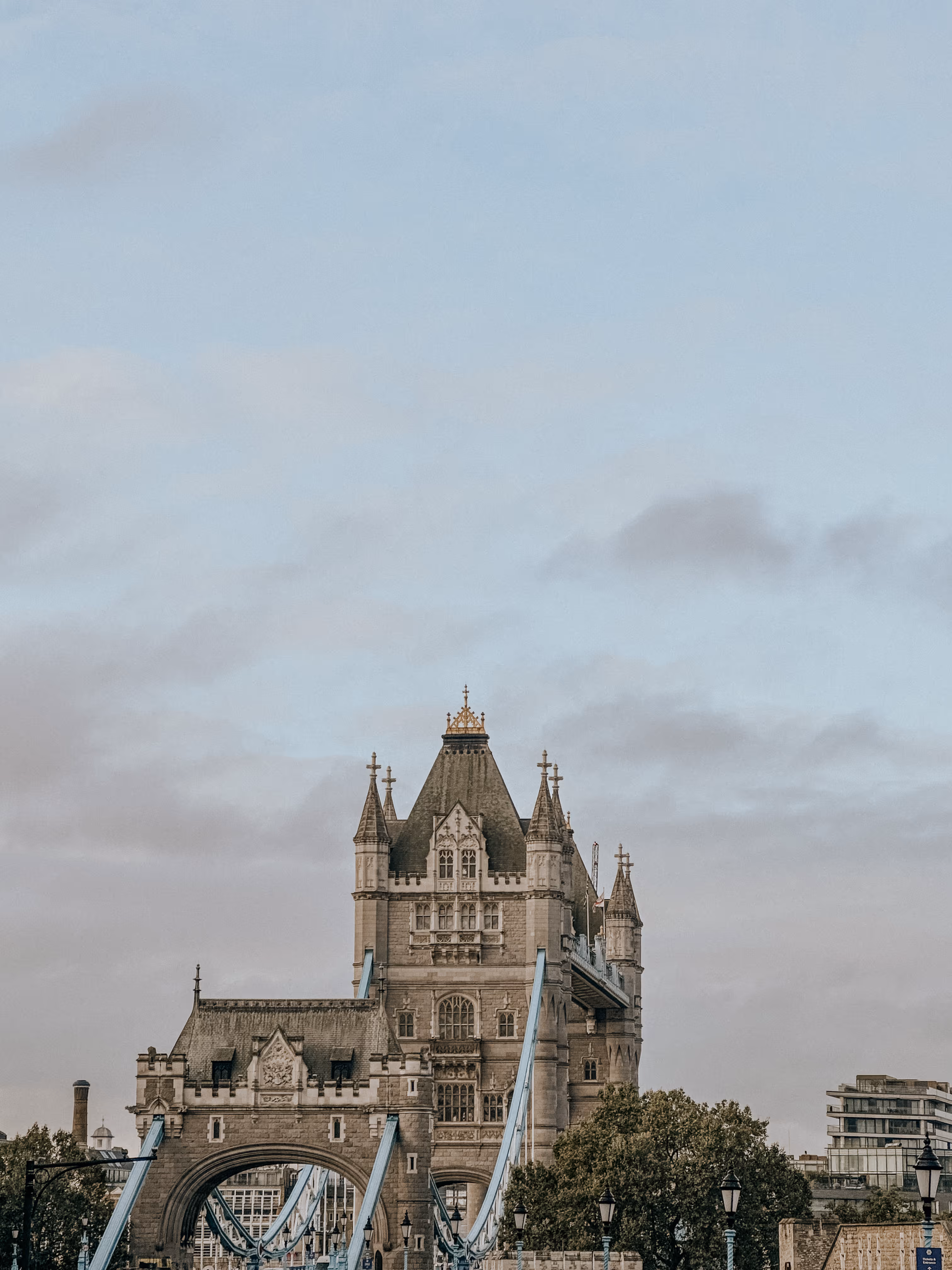 Tower Bridge in London with stone towers and blue suspension cables under a cloudy sky.