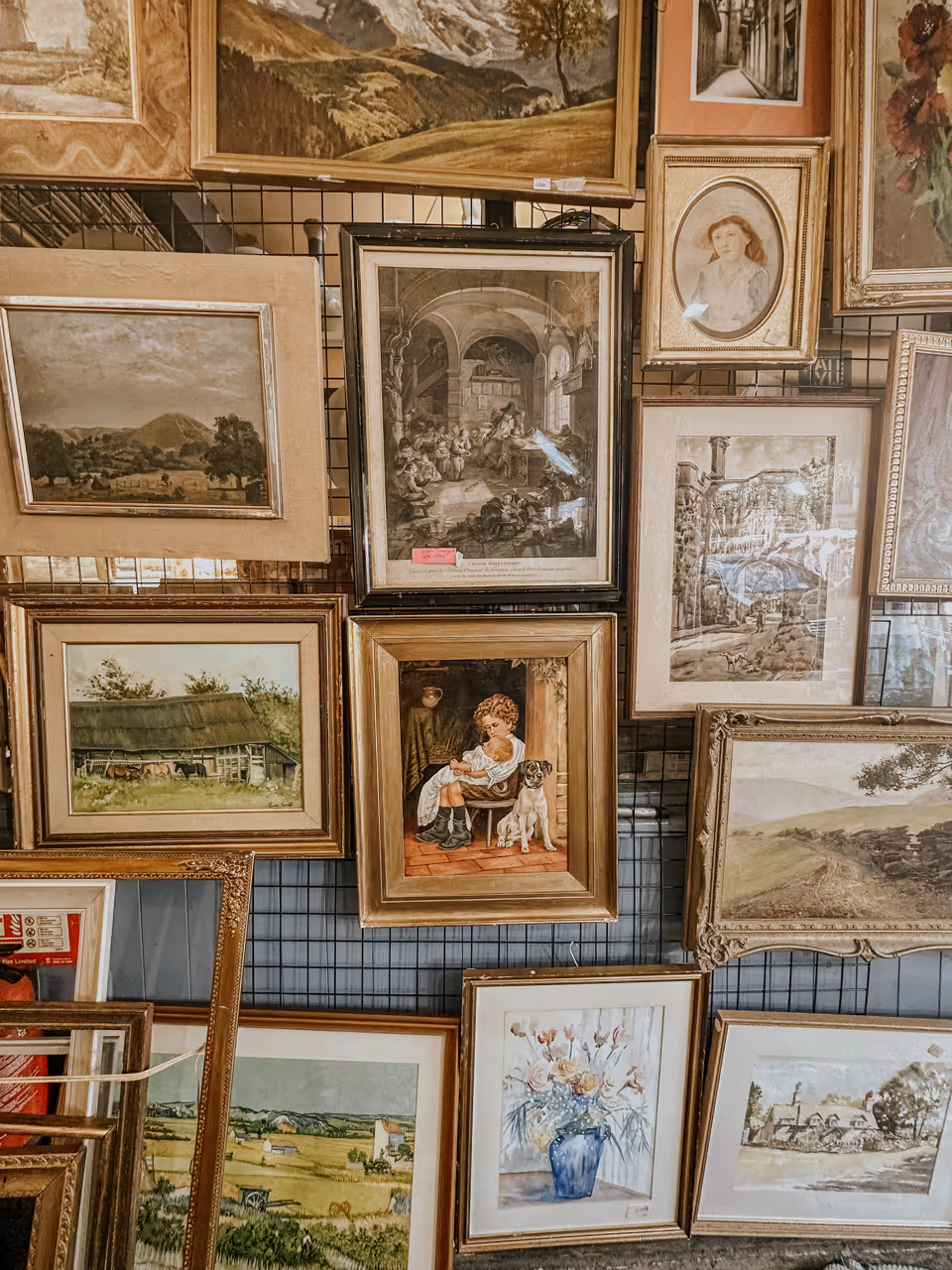 Wall display of various framed paintings including landscapes, a child with a dog, flowers, and portraits.