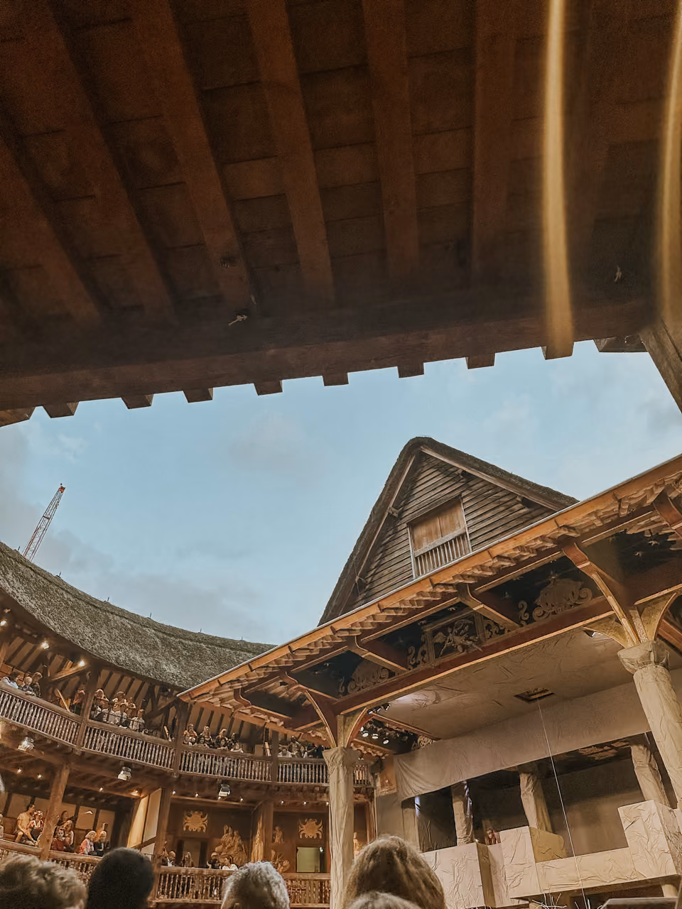 Audience watching a play in a historic open-air wooden theatre with thatched roof under sky.