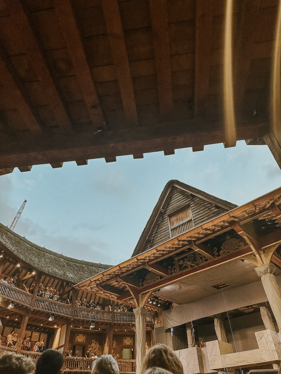Audience watching a play in a historic open-air wooden theatre with thatched roof under sky.
