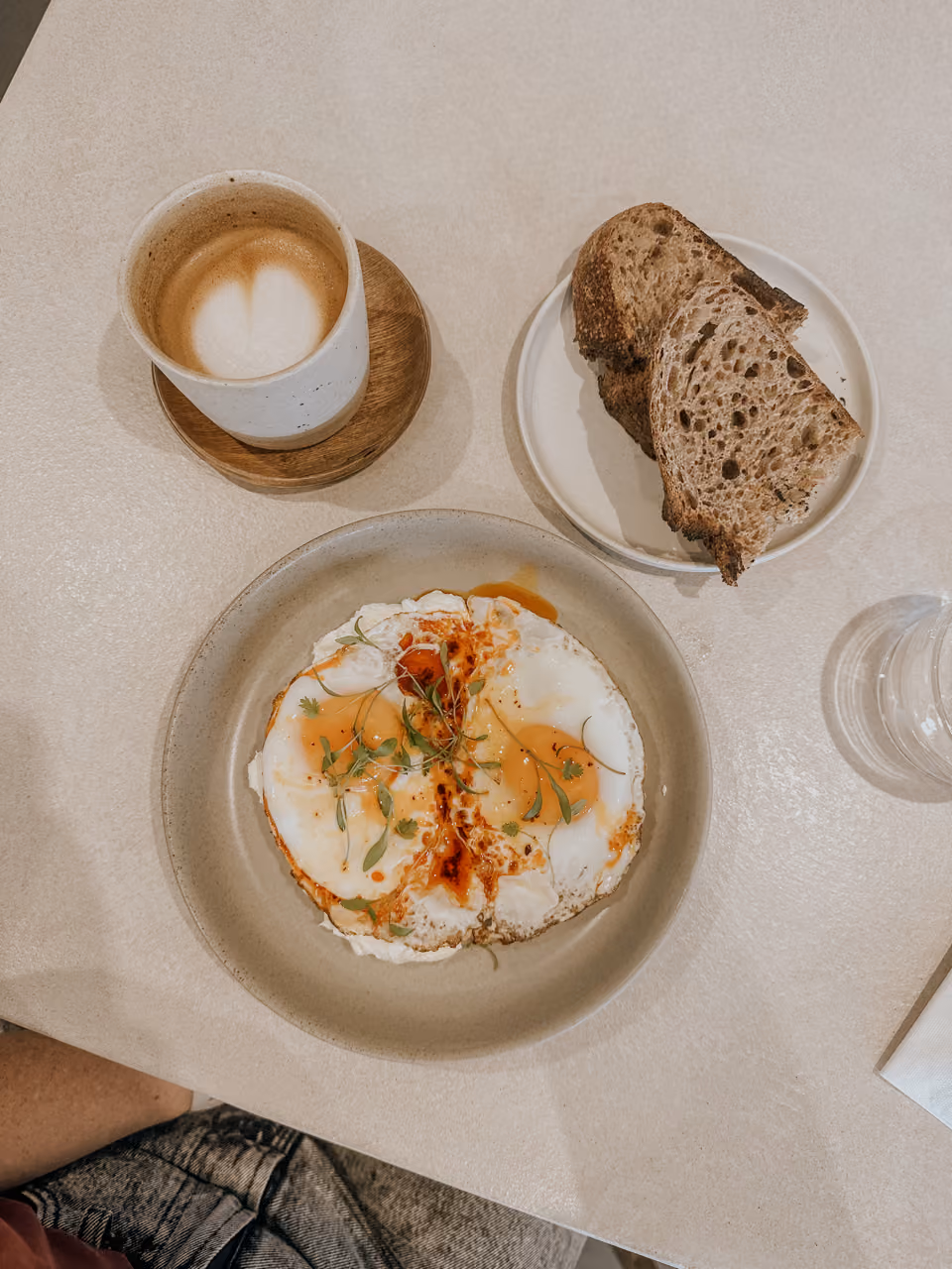 Two fried eggs topped with greens on a plate, served with two slices of bread and a latte in a ceramic cup.