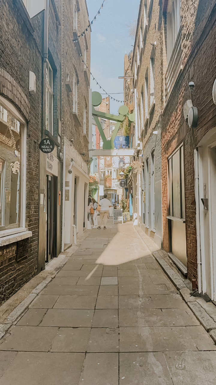 Narrow pedestrian alley with brick buildings, string lights, and people walking in daylight.