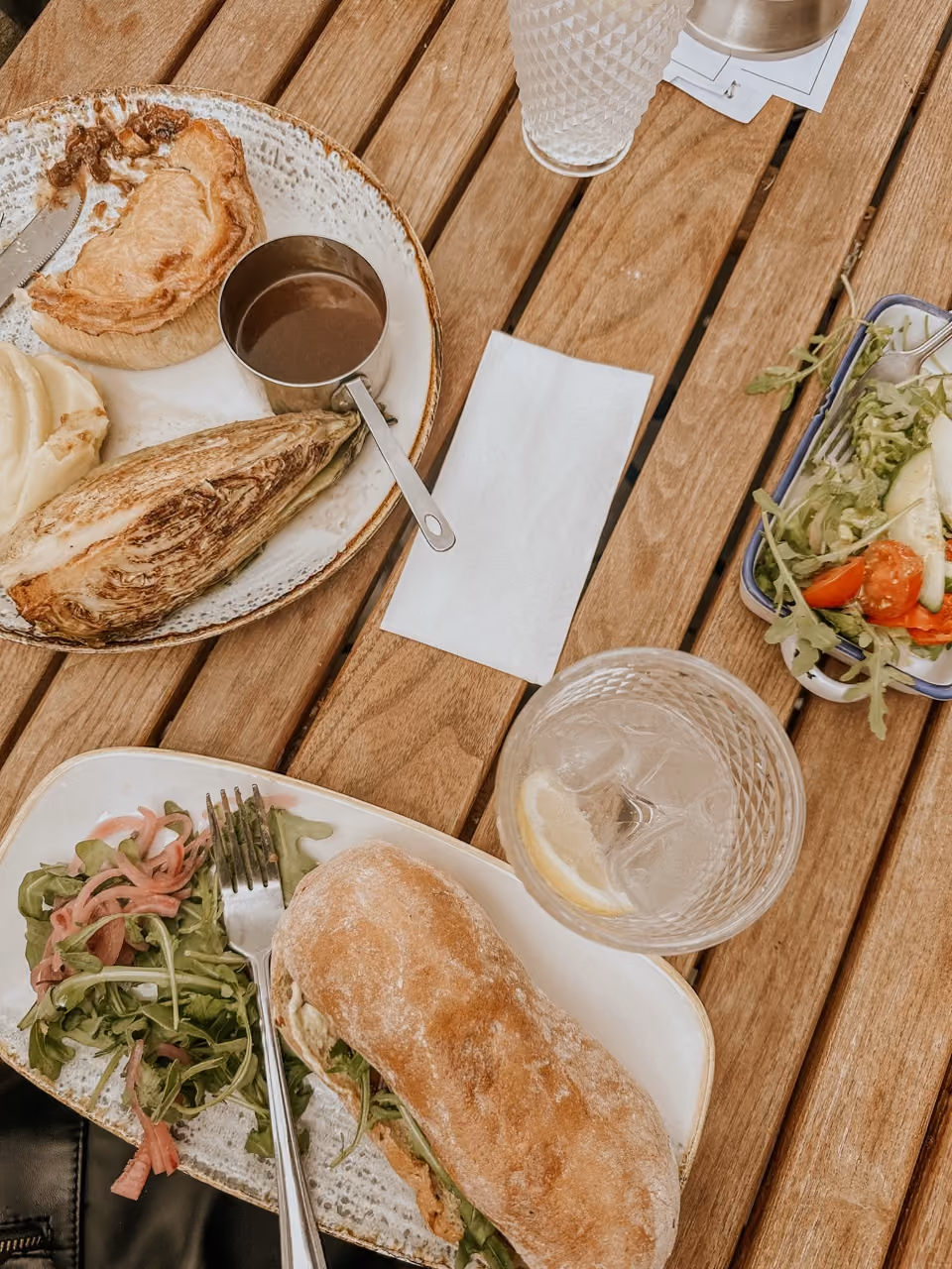 Wooden table with plates of sandwich, salad, gravy, pastry, and glasses with drinks and lemon slices.