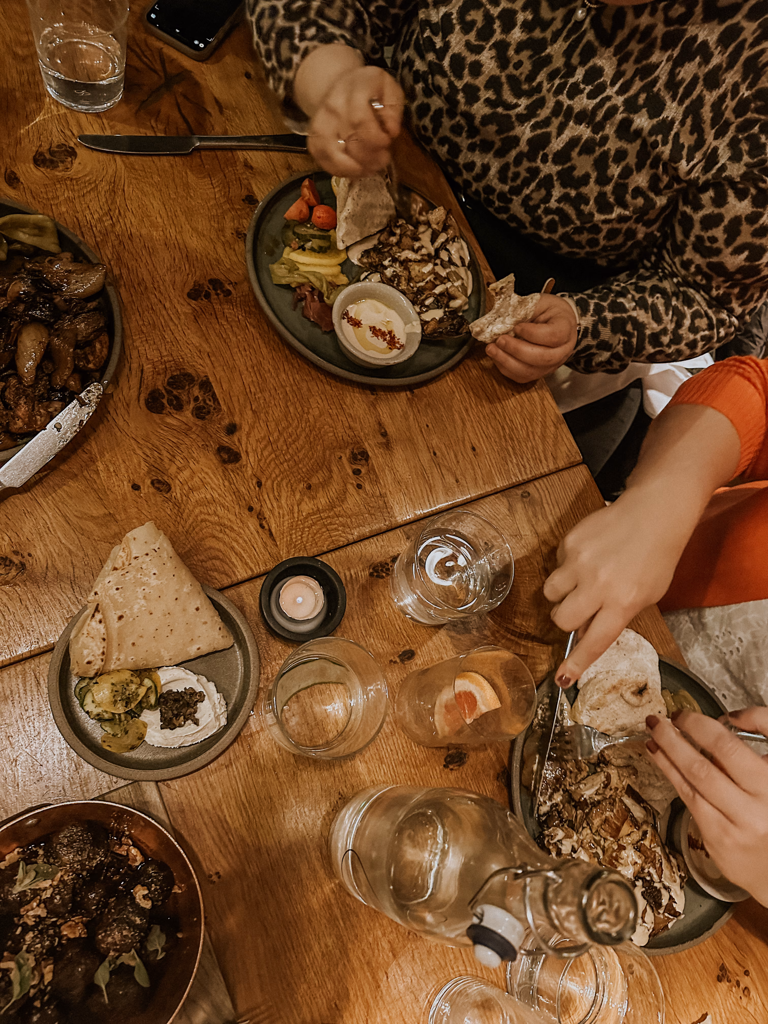 Two people eating a meal with grilled chicken, pita bread, vegetables, and dips on a wooden table.