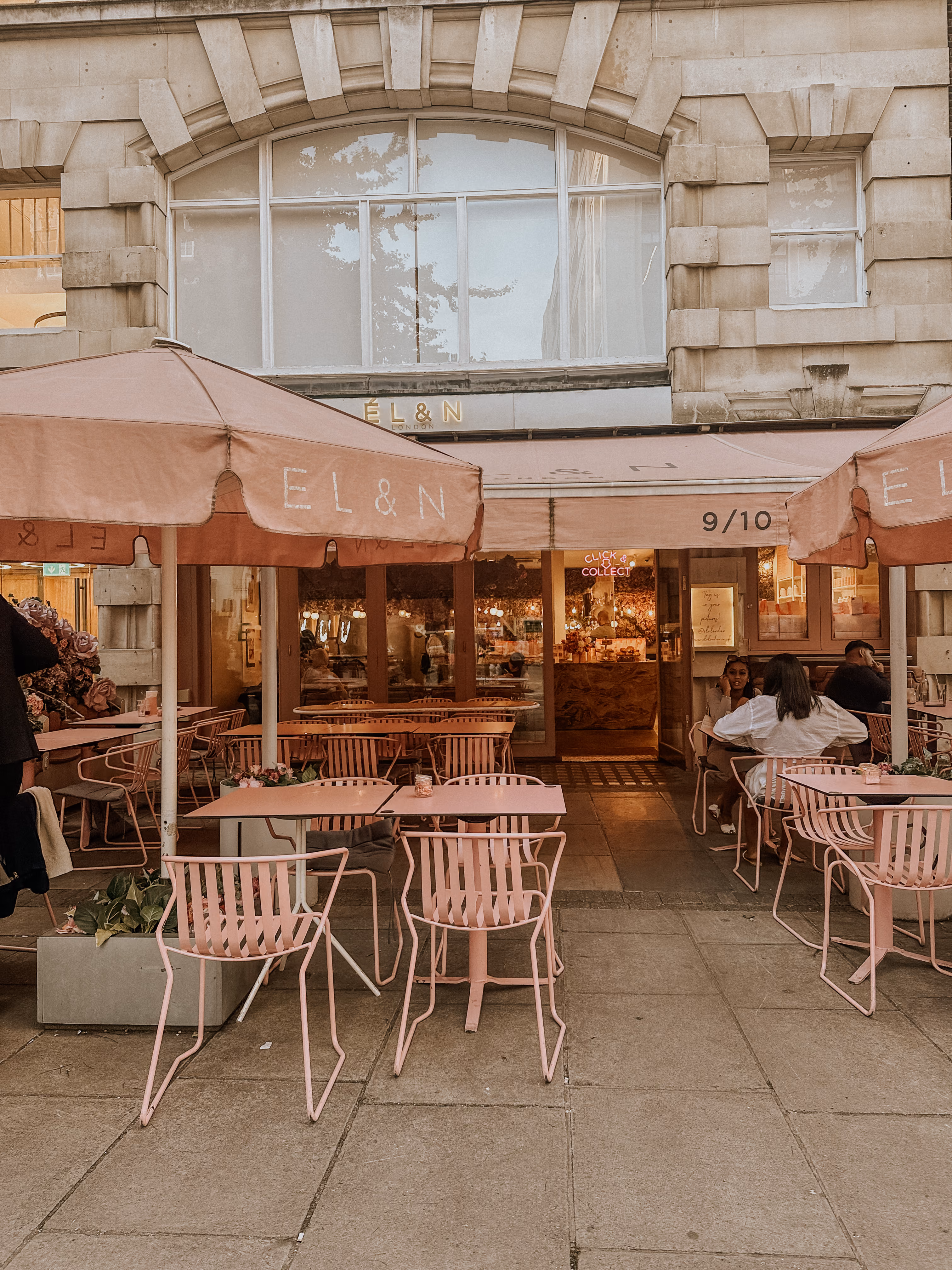 Outdoor café with pink tables, chairs, umbrellas, and people seated under an awning.