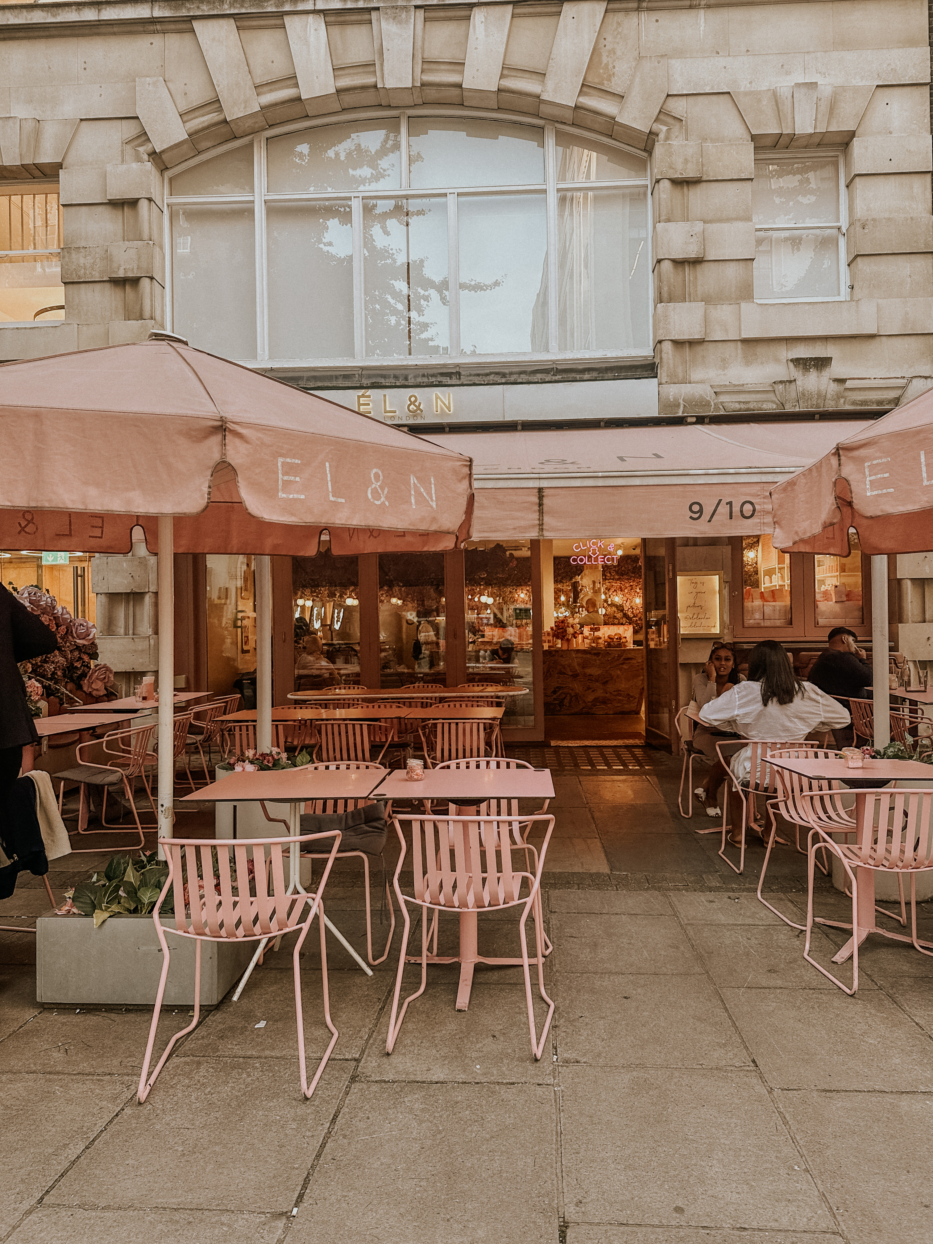 Outdoor café with pink tables, chairs, umbrellas, and people seated under an awning.