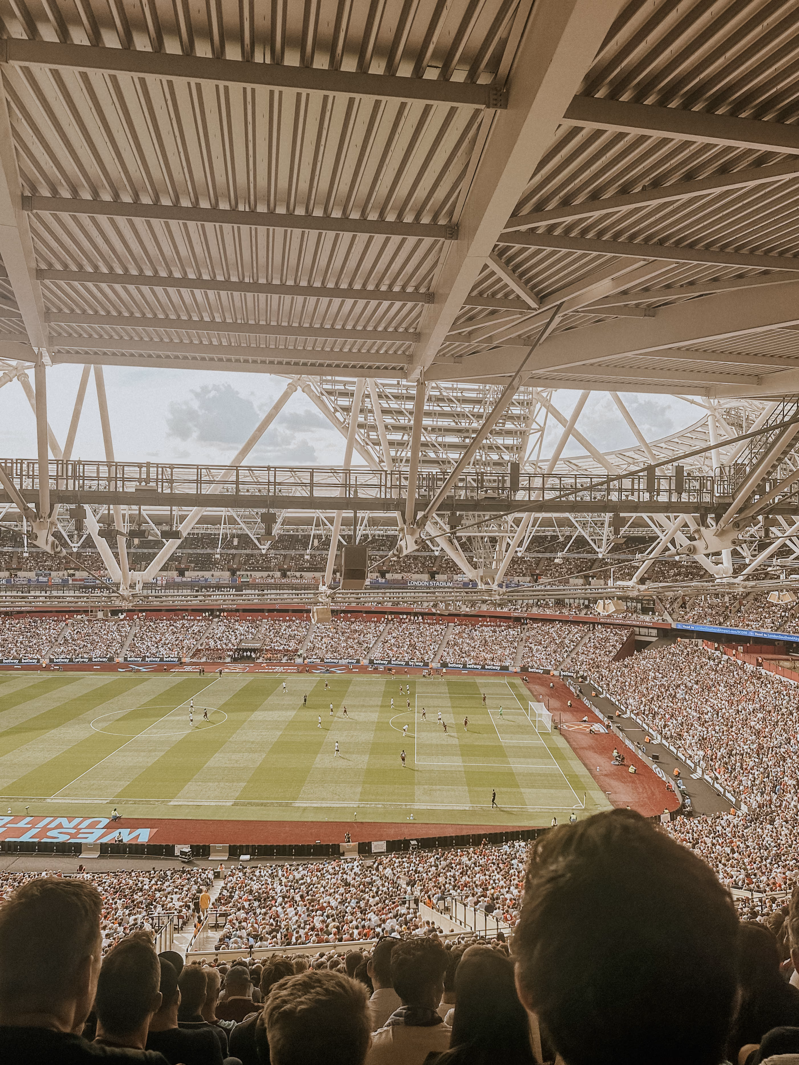 Crowded stadium with fans watching a soccer match under a large metal roof structure.