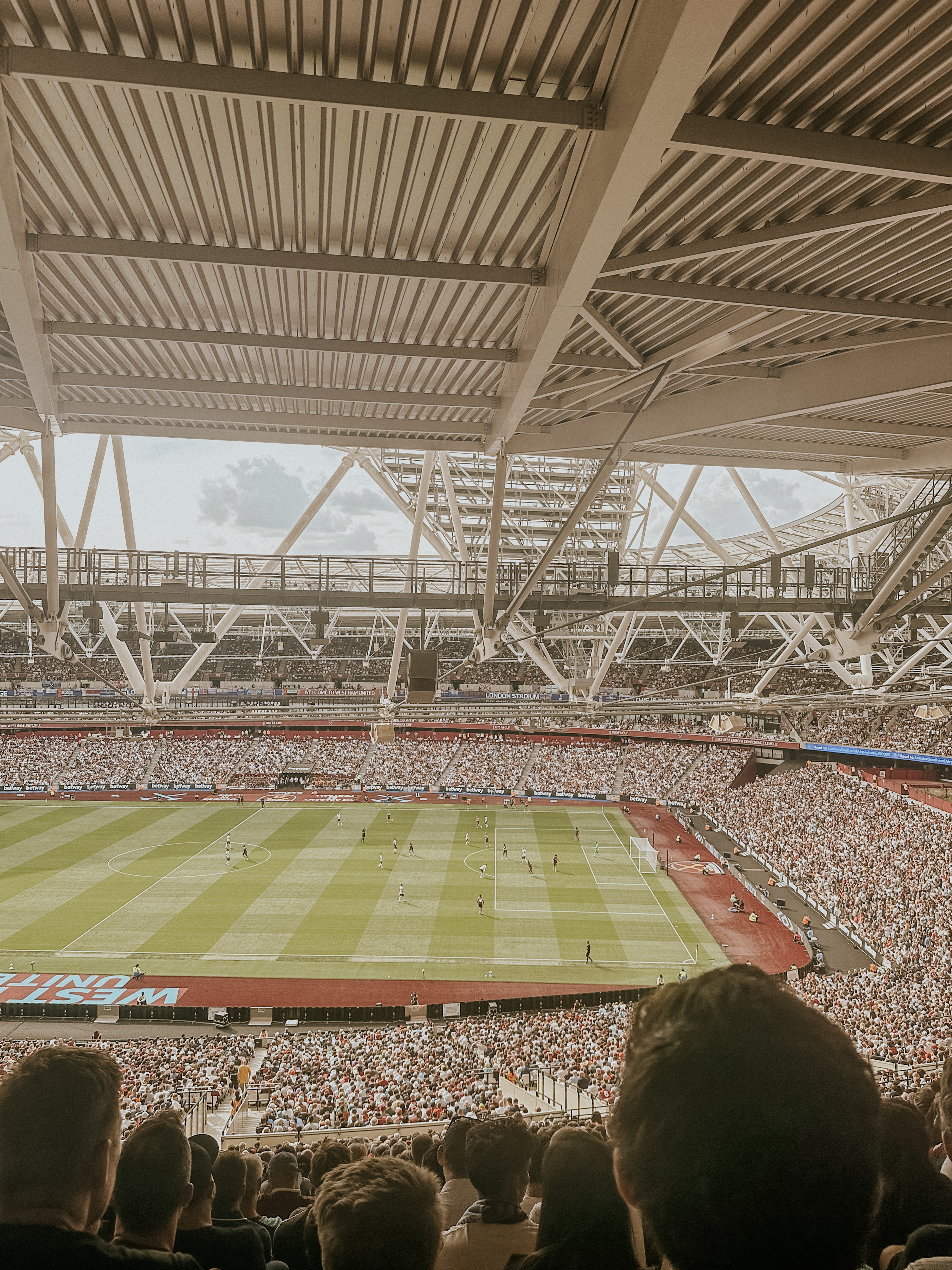 Crowded stadium with fans watching a soccer match under a large metal roof structure.