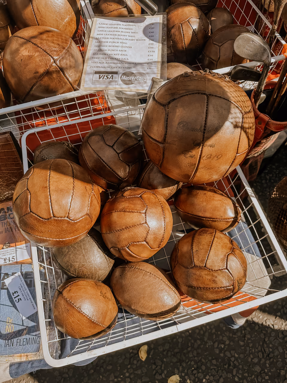 Basket filled with vintage brown leather footballs and rugby balls for sale at a market stall.