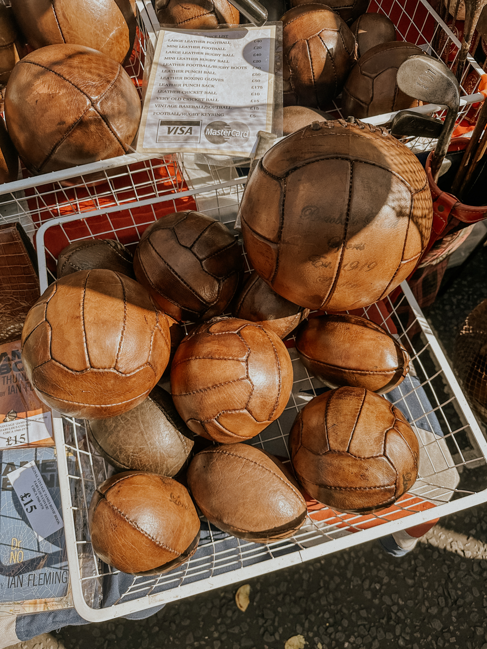 Basket filled with vintage brown leather footballs and rugby balls for sale at a market stall.