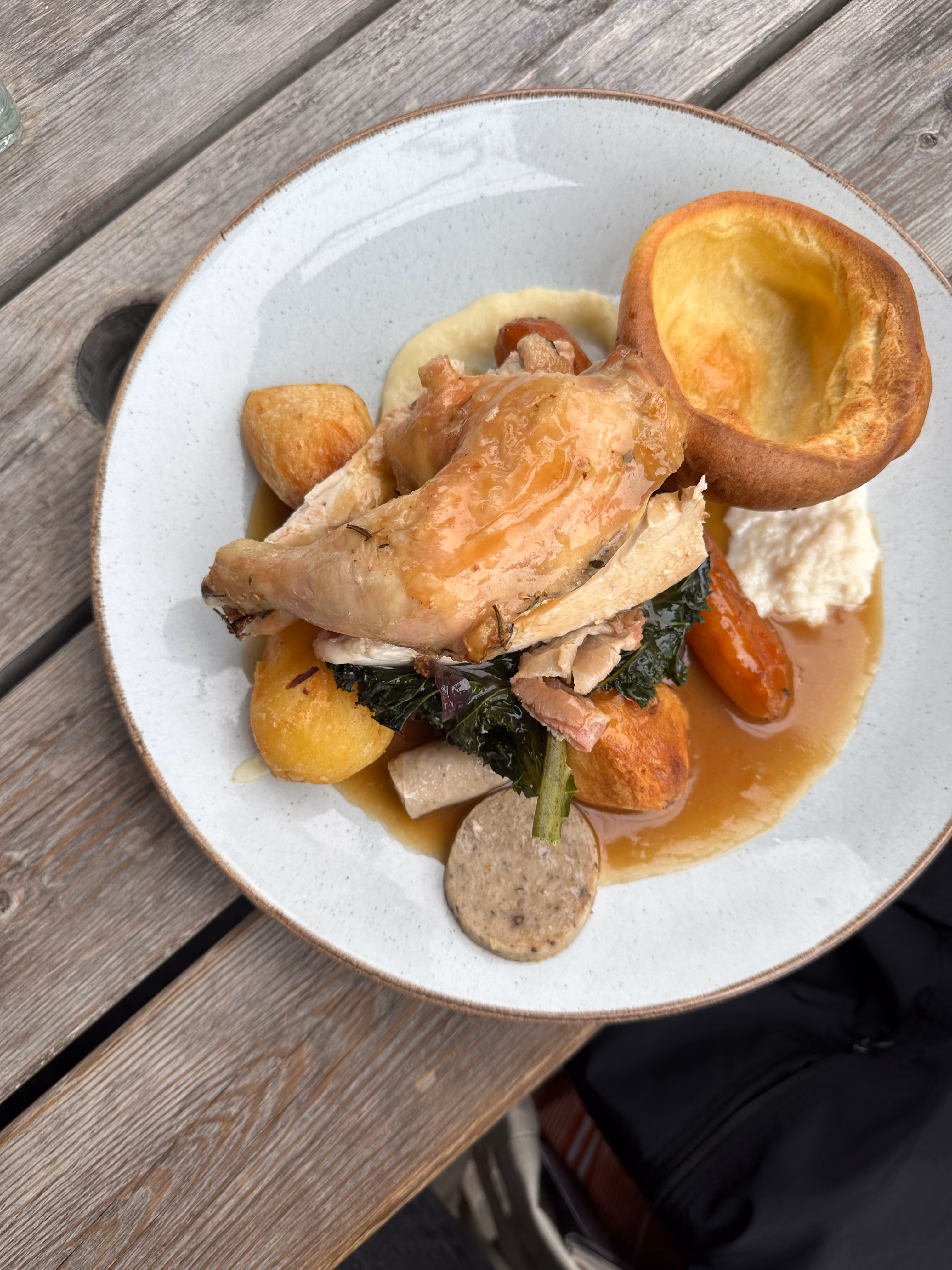 Plate with roasted chicken, Yorkshire pudding, roasted potatoes, vegetables, sausage, and gravy on a wooden table.