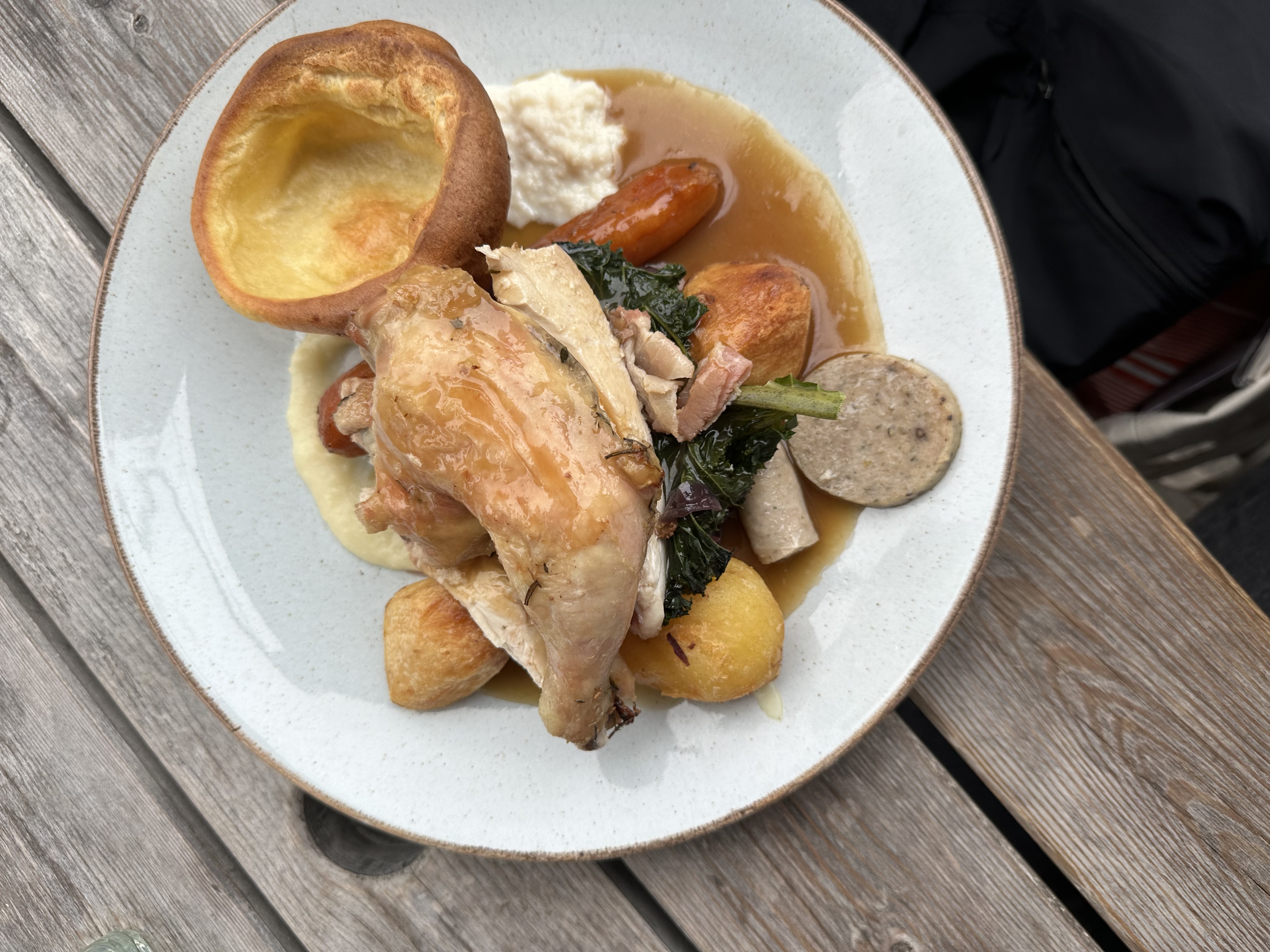 Plate with roasted chicken, Yorkshire pudding, roasted potatoes, vegetables, sausage, and gravy on a wooden table.