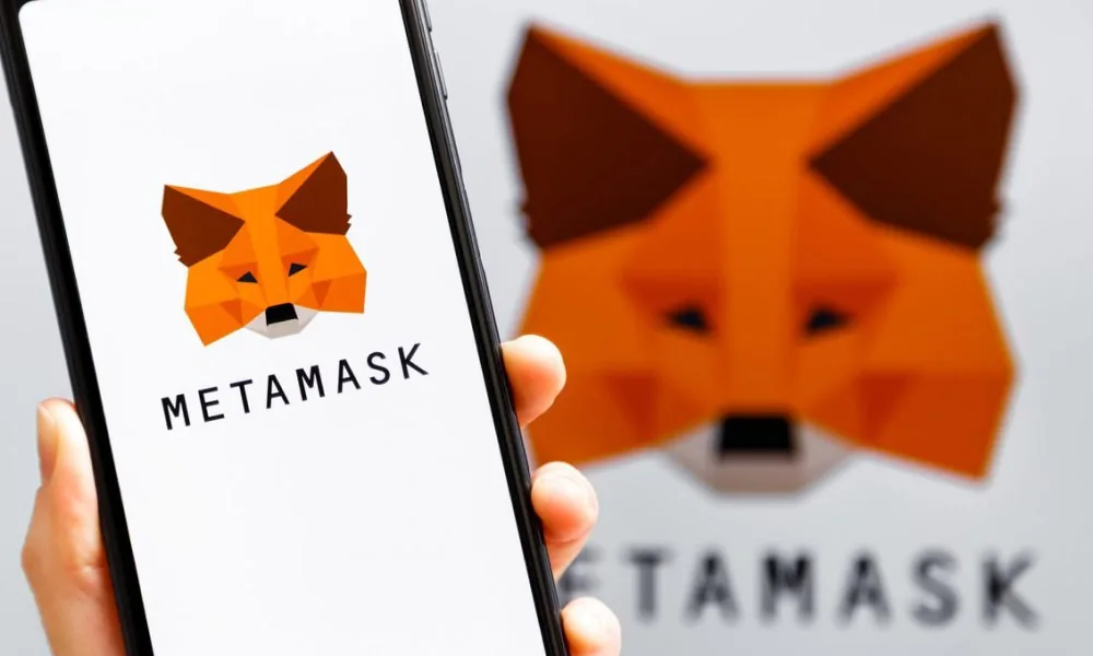 Hand holding a phone showing the MetaMask app in front of a large MetaMask fox logo